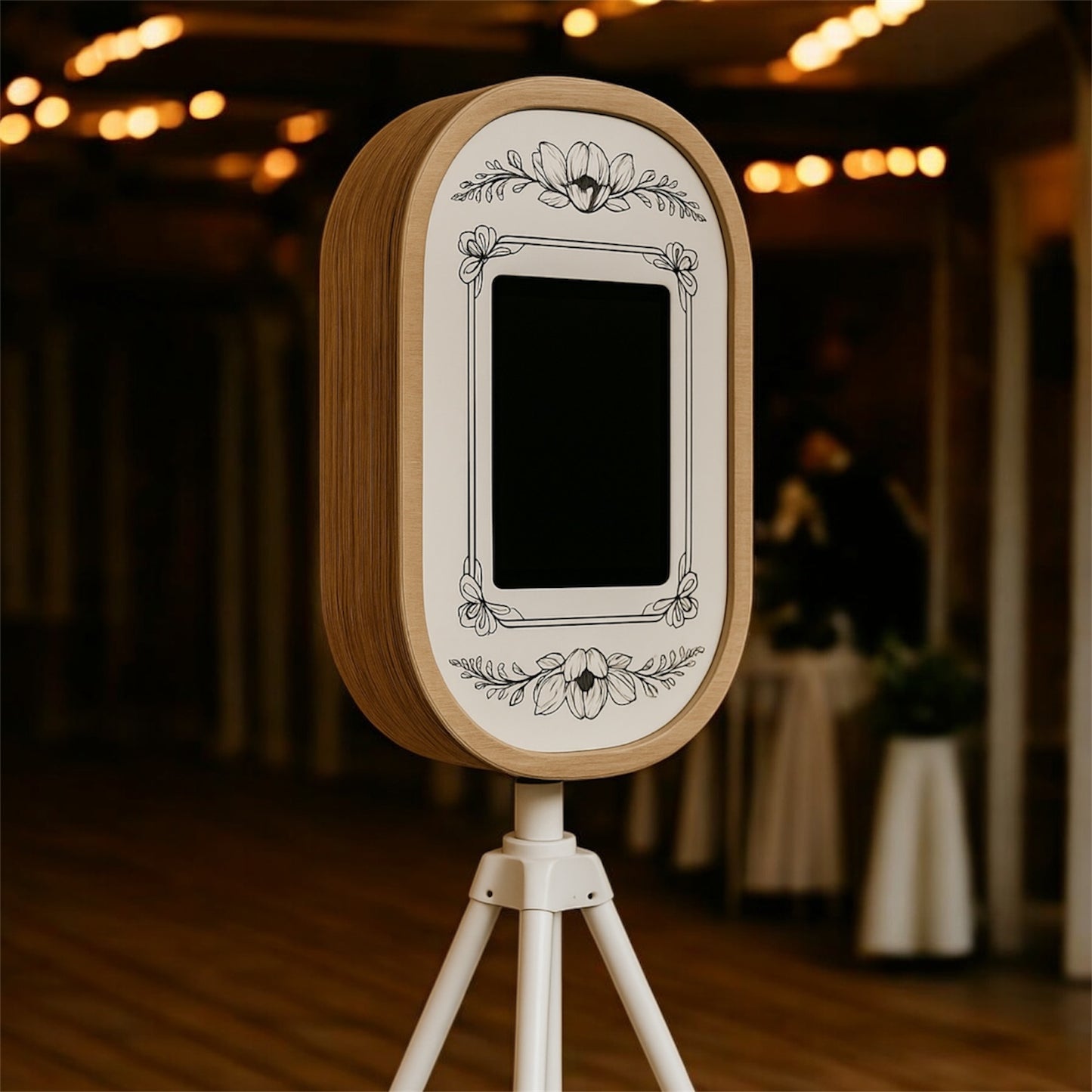 Handcrafted rustic photo booth with floral engraving on a wooden frame, mounted on a white tripod and styled for a wedding venue.
