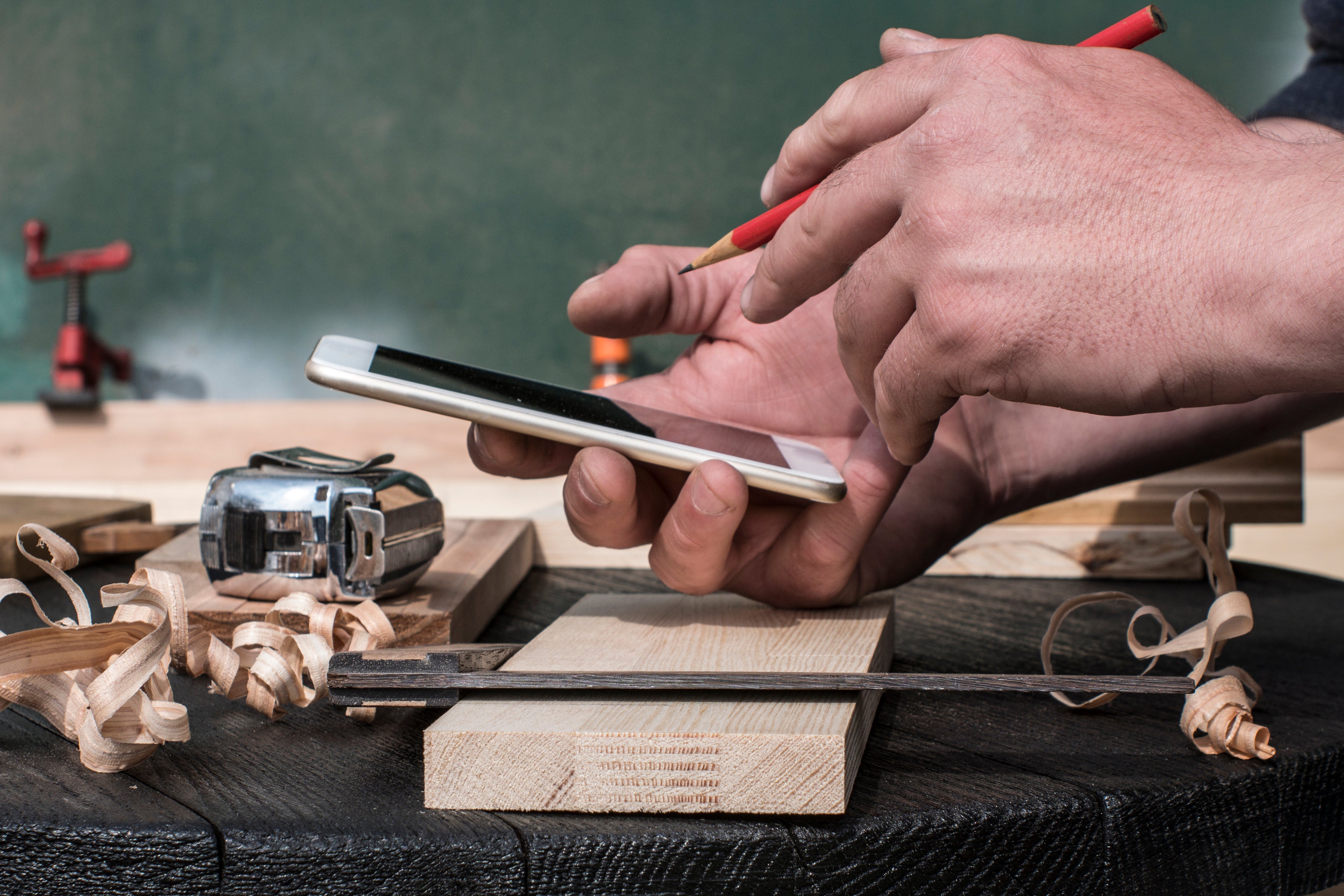 Close-up of a woodworker using a smartphone at a workshop desk, surrounded by wood shavings and tools.