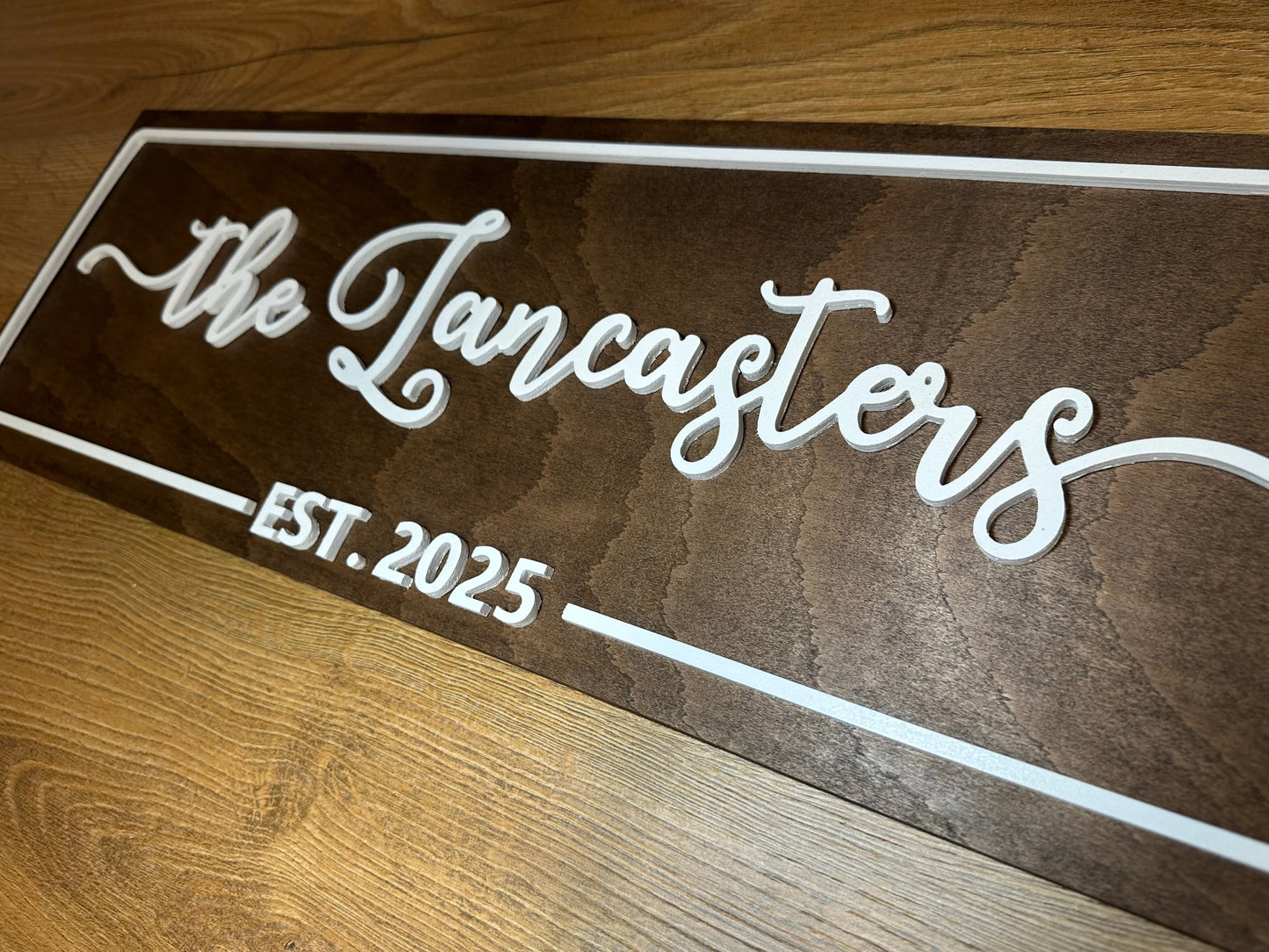 Close-up of a custom wooden family name sign with raised white lettering showing “The Lancasters – Est. 2025” on a woodgrain surface.
