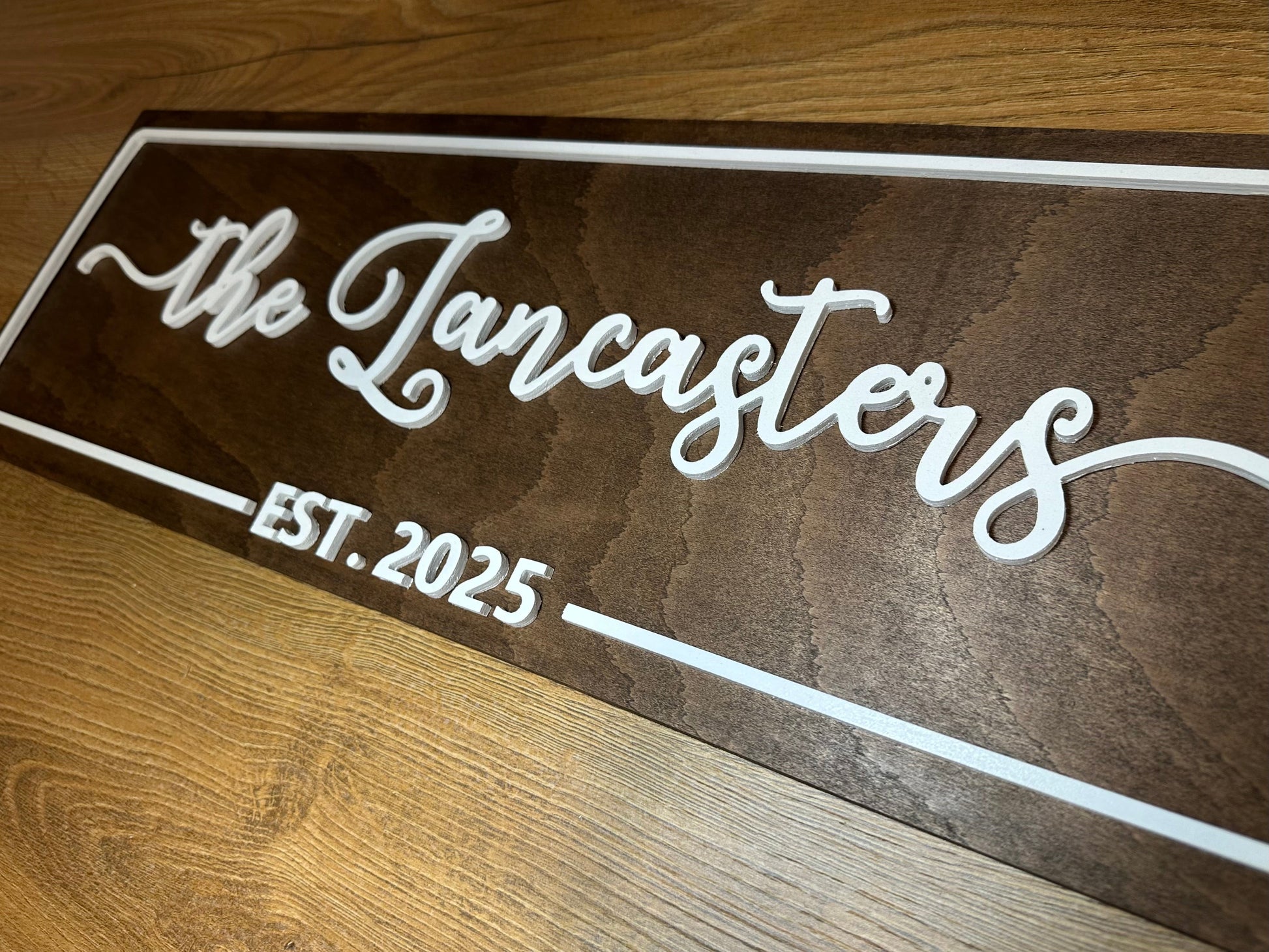 Close-up of a custom wooden family name sign with raised white lettering showing “The Lancasters – Est. 2025” on a woodgrain surface.