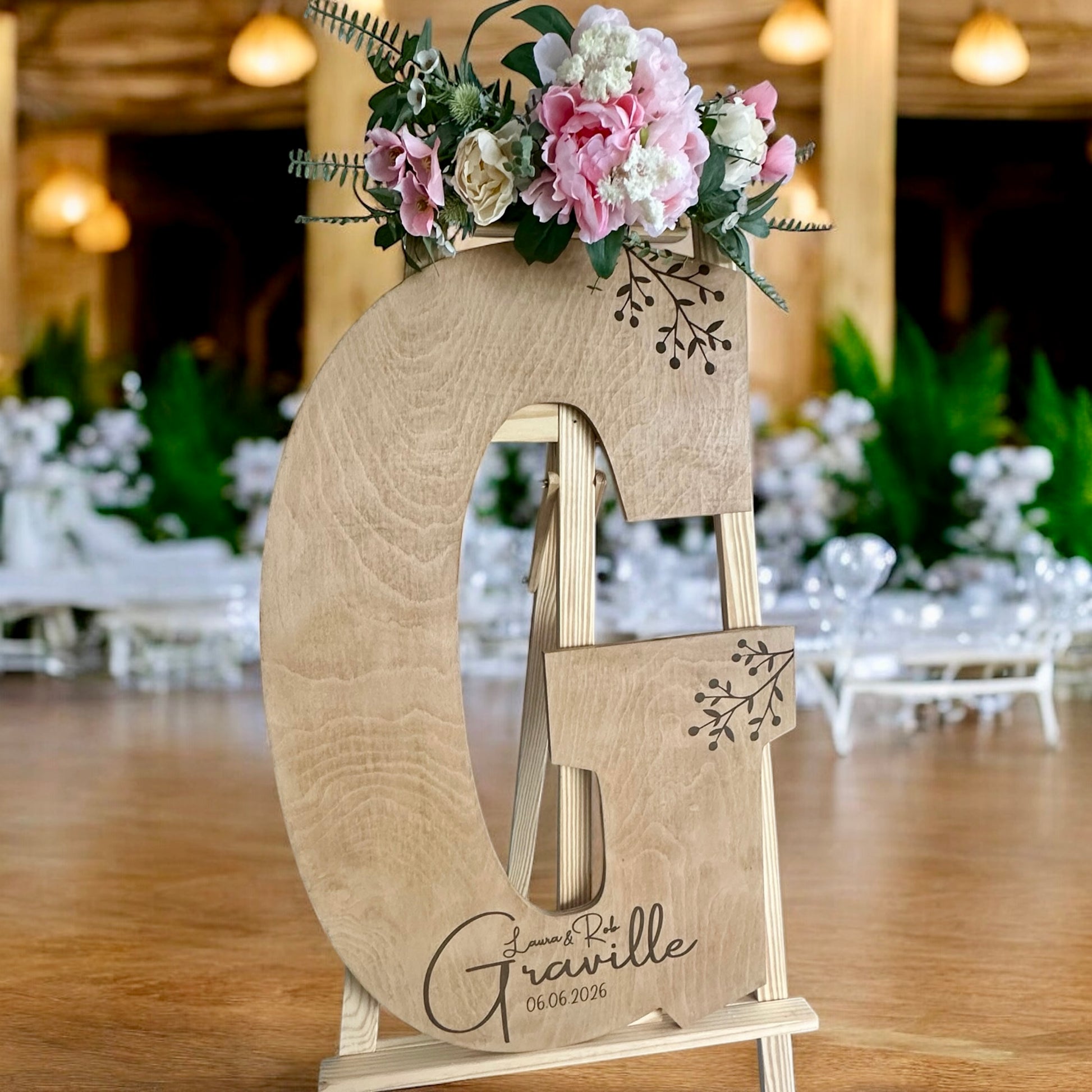 Wooden guestbook letter engraved with “Laura & Rob Graville – 06.06.2026” and decorative berry branch detail, placed on an easel.