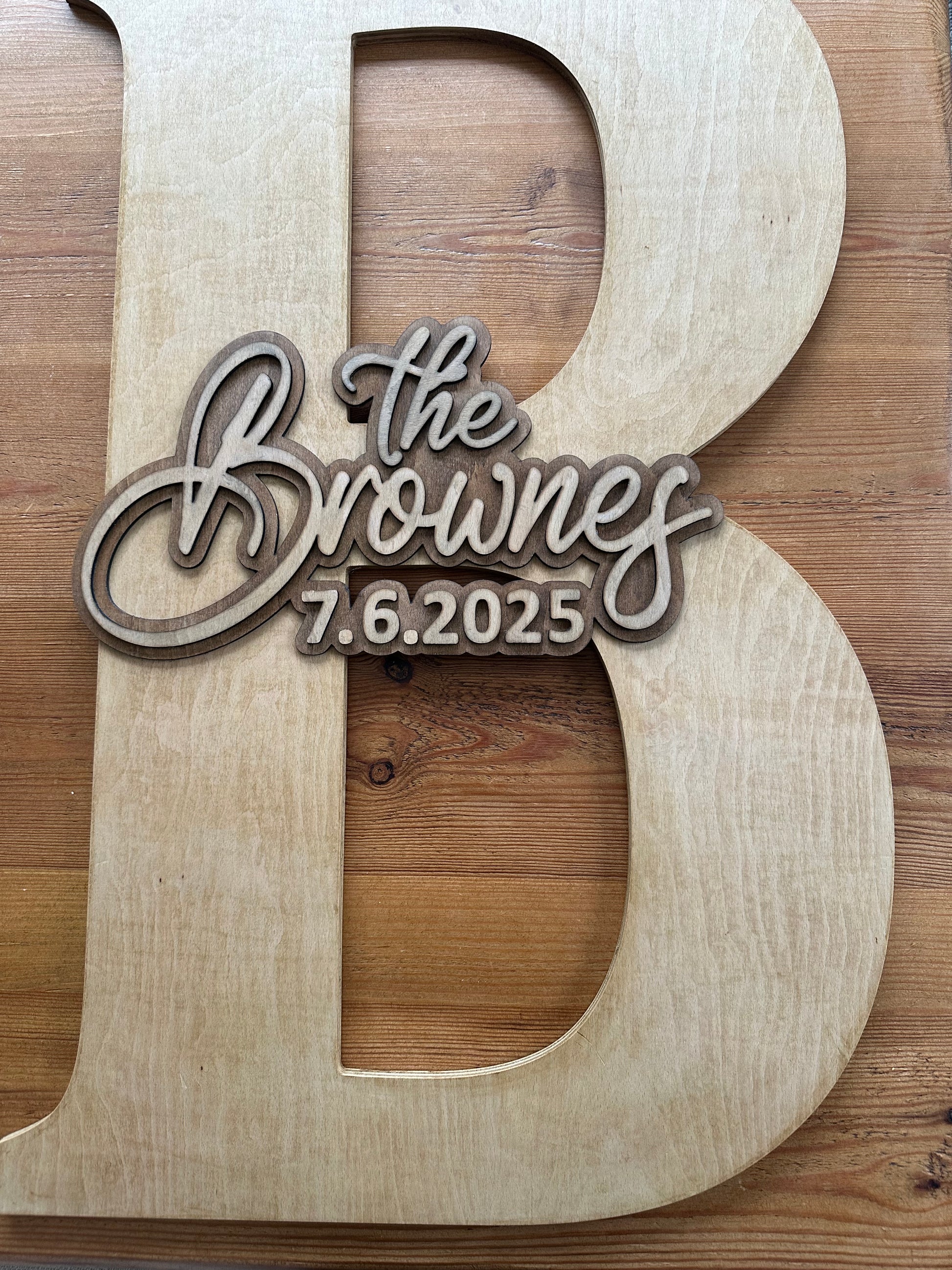 Flatlay image of wooden guestbook letter “B” engraved with “The Brownes – 7.6.2025” placed on a rustic wooden table.