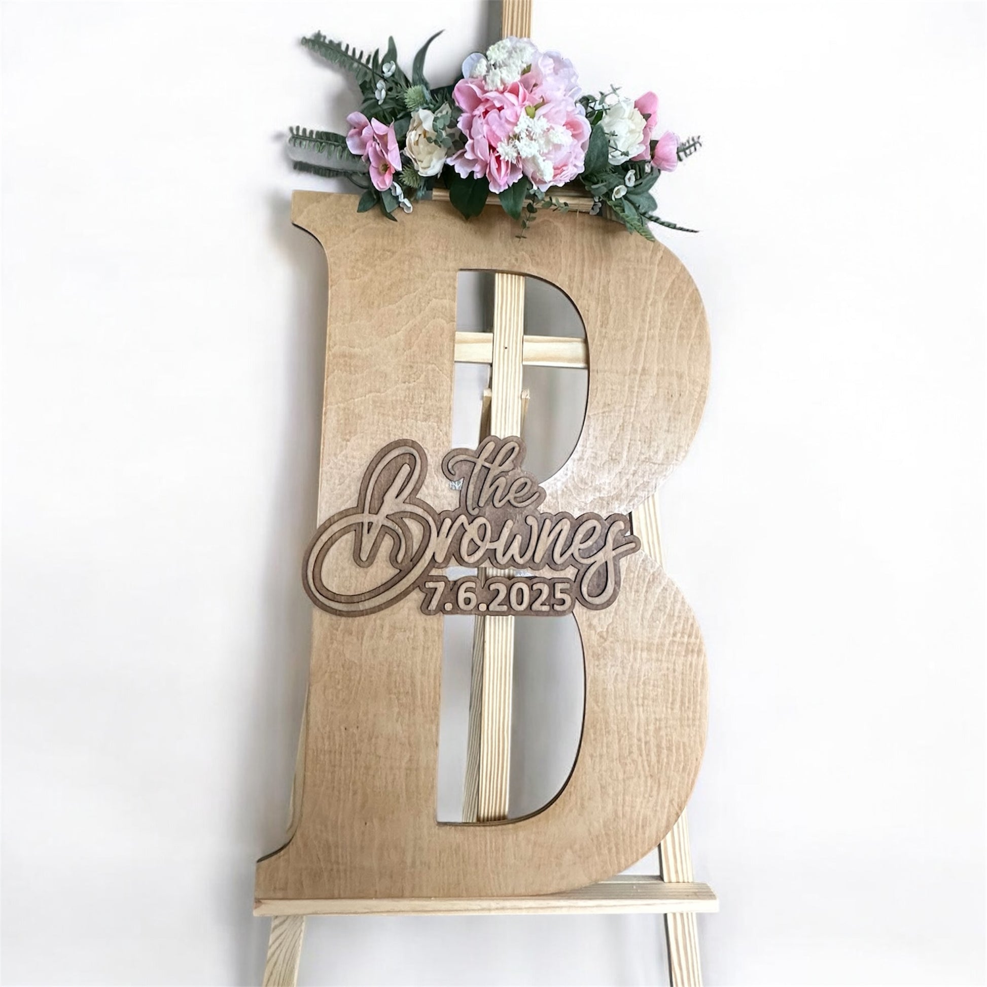Large personalised wooden guestbook letter “B” with “The Brownes – 7.6.2025” engraved, displayed on an easel with pink and white flowers.