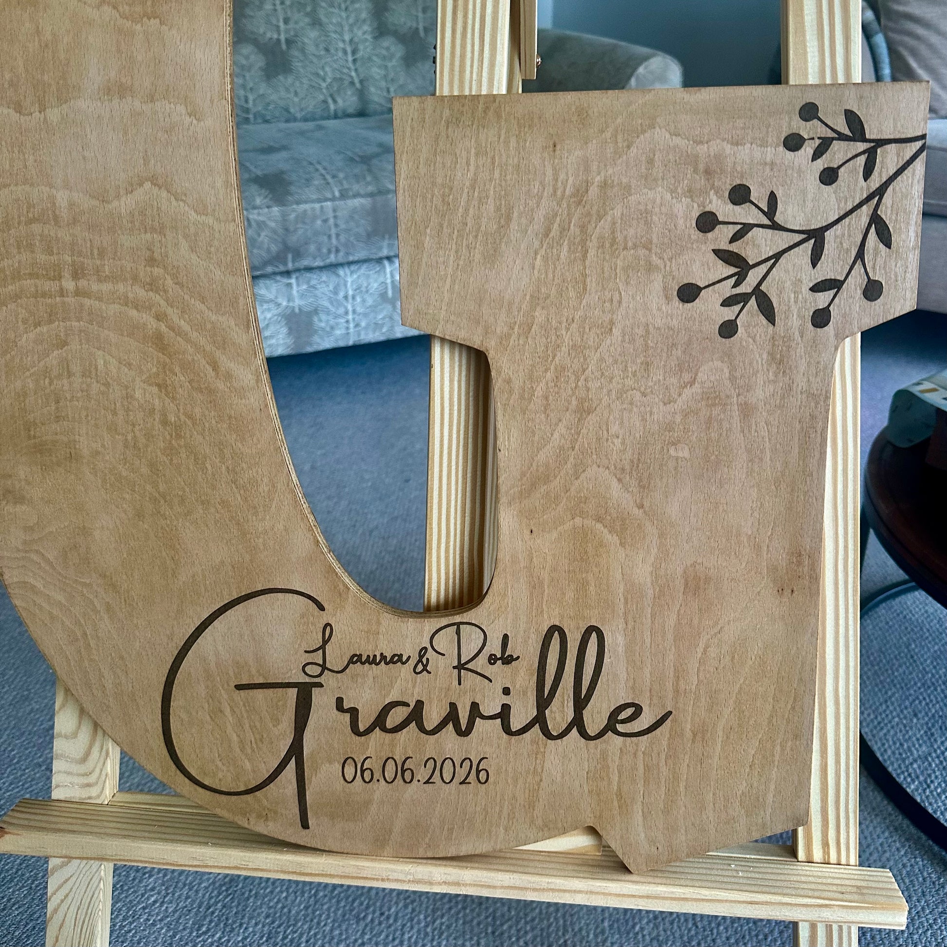 Large wooden guestbook letter “G” on an easel with floral top piece, engraved with “Laura & Rob Graville – 06.06.2026” and botanical accents.
