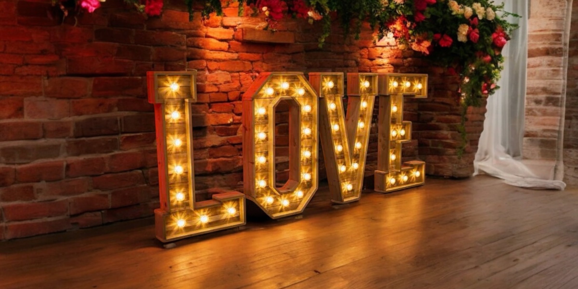 Large rustic LOVE sign lit with warm bulbs, set against a red brick wall and accented with flowers and draped fabric.