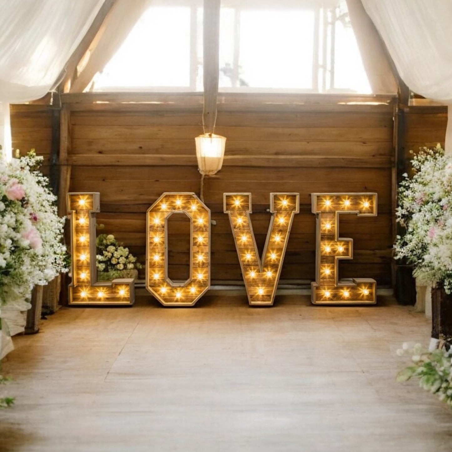 Illuminated LOVE letters in a rustic barn aisle, surrounded by flowers and draped curtains.