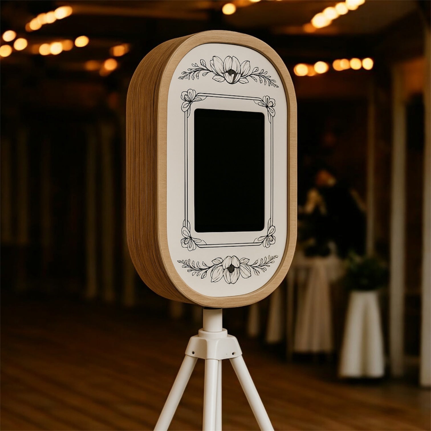Handcrafted rustic photo booth with floral engraving on a wooden frame, mounted on a white tripod and styled for a wedding venue.