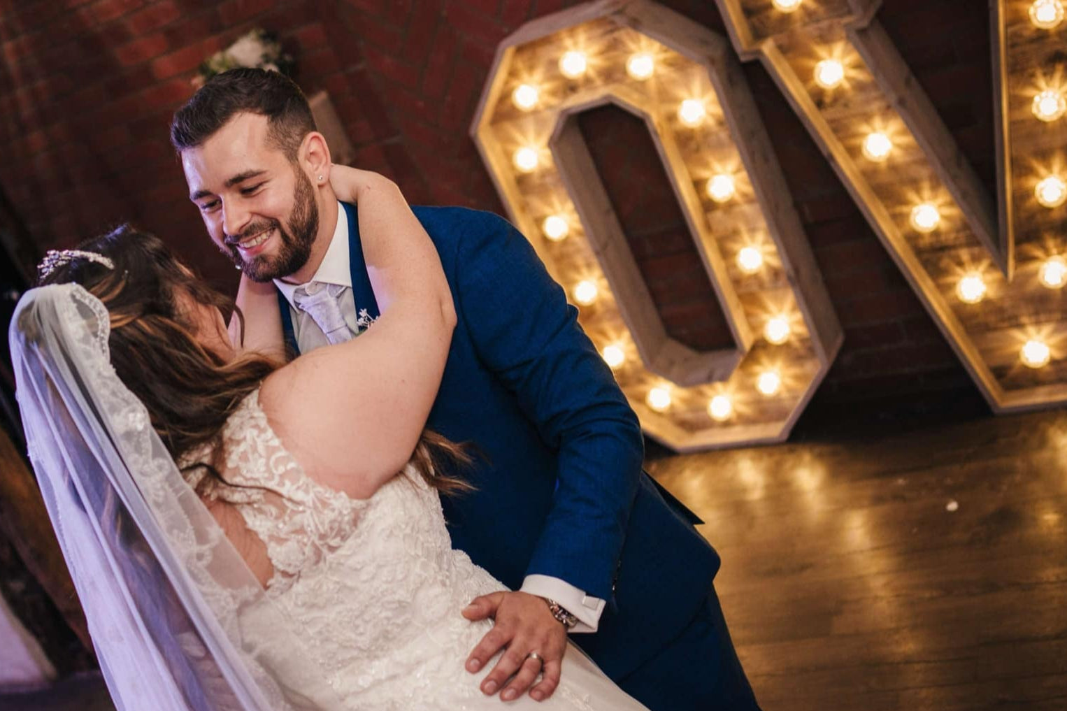 Bride and groom share a romantic first dance with large illuminated wooden LOVE letters glowing warmly in the background.